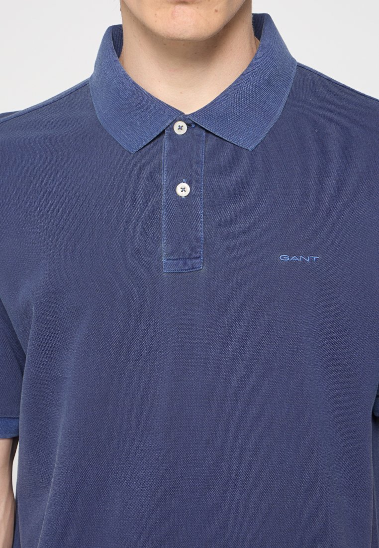 Mężczyzna w granatowej koszulce polo GANT z dwoma białymi guzikami zapiętymi pod kołnierzykiem.
