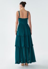 Abito lungo a balze color blu-verde con spalline sottili regolabili, orlo arricciato e corpetto arricciato. Tessuto trasparente e leggero, silhouette fluente.