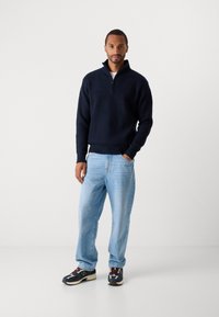 Mężczyzna w granatowym swetrze typu quarter-zip, jasnoniebieskich dżinsach i wielokolorowych sneakersach, stojący z jedną ręką w kieszeni na jednolitym tle.