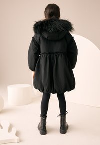 Black puffer jacket with a faux fur hood, elastic waist, and balloon shape. Worn over black tights and sturdy black ankle boots.