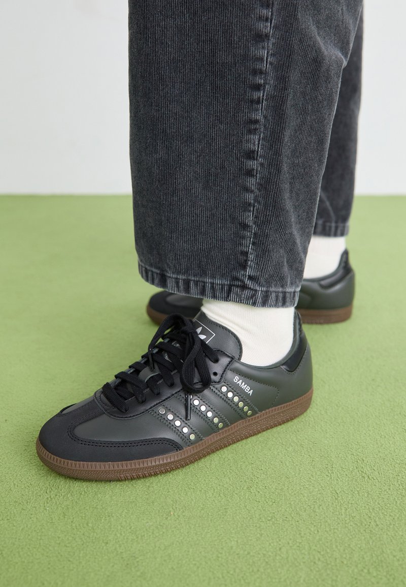 Black leather sneakers with perforated stripes and brown rubber sole, paired with grey corduroy pants and white socks on a green surface.