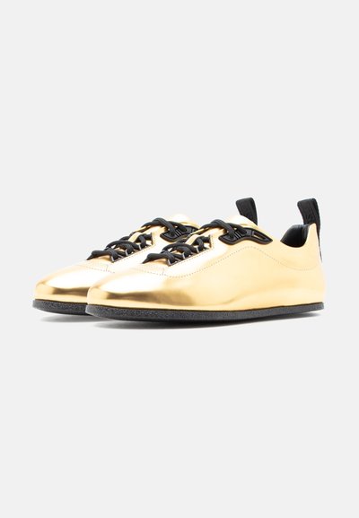 Pár lesklých zlatých tenisiek s čiernymi šnúrkami, čiernymi ťahacími pútkami a textúrovanými čiernymi podrážkami na jednoduchom svetlom pozadí.