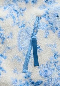 Hellblauer Reißverschluss, teilweise geöffnet, auf weißem und blauem, flauschigem Fleece-Stoff mit abstraktem Muster.