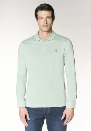 Uomo con indosso una polo verde chiaro a maniche lunghe con tre bottoni e jeans blu, in piedi davanti a uno sfondo semplice.