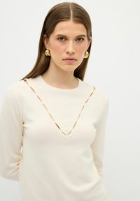 Cremfarbener Strickpullover mit V-Ausschnitt und Aussparungen am Hals. Lange Ärmel und eine glatte Textur. Akzentuierte goldene Creolen.