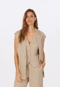 Beige ärmelloser Blazer mit Revers und drei vorderen Knöpfen, kombiniert mit einer passenden Weste. Der Stoff wirkt glatt mit minimaler Struktur.