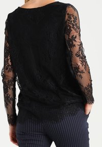 Blusa nera in pizzo con maniche lunghe trasparenti, caratterizzata da motivi floreali e orlo a scallop. Abbinata a pantaloni a righe.