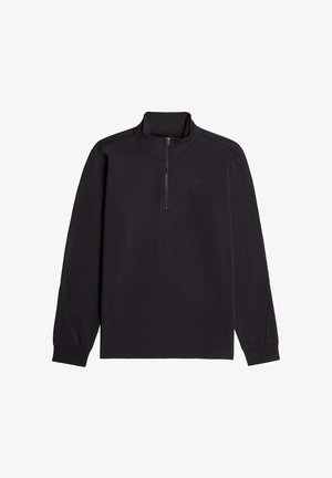 Homme portant une chemise noire à manches longues et un pantalon clair, vu de dos sur un fond uni.