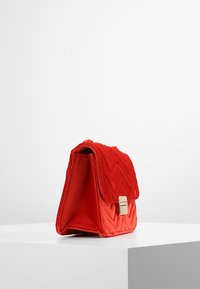 Červená prošívaná sametová kabelka s klopou, zlatými kovovými prvky a pevnou strukturou, která má šikmé vzory a měkkou texturu.