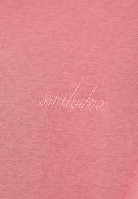 Ροζ ύφασμα με λεπτή κεντητική της λέξης "smilodox" με καλλιγραφική ραφή.