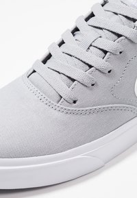 Baskets en toile grises avec un dessus texturé, lacets plats et semelle en caoutchouc blanc. Présente un logo distinctif sur le côté et des détails de couture.