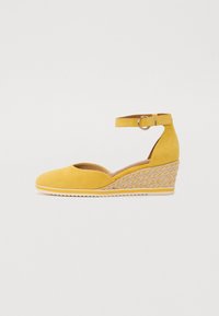 Chaussure compensée en daim jaune avec une bride au niveau de la cheville, un détail en métal circulaire et une texture tissée sur le talon compensé. Design à bout rond.
