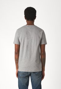 Γκρίζο κοντομάνικο μπλουζάκι από βαμβακερό μείγμα, με στρογγυλή λαιμόκοψη και λεία υφή. Φοριέται με μπλε τζιν, με θέα από πίσω και τατουάζ.