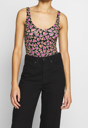 Kvinde iført en ærmeløs sort top med pink og gule hjerteformer kombineret med højtsiddende sorte bukser mod en ensartet baggrund.