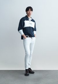 Giovane uomo in piedi con le mani in tasca, indossa una maglia a maniche lunghe blu navy e bianca, pantaloni bianchi e stivali marrone scuro su un pavimento grigio.