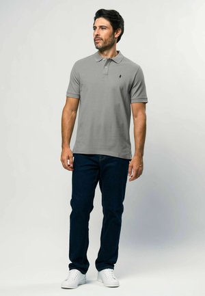 Ember áll, szürke rövid ujjú galléros pólót, sötétkék farmert és fehér tornacipőt visel, oldalra néz egy egyszínű háttér előtt.