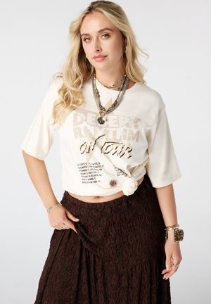 Jonge vrouw met lang blond haar draagt een geknoopte witte "Desert Rhythm On Tour" t-shirt, bruine rok, gelaagde kettingen en armbanden.