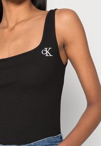 Svart ribbad linne med rundad halsringning, prytt med en vit "ck"-logotyp på vänster bröst, tillsammans med blå denimjeans.