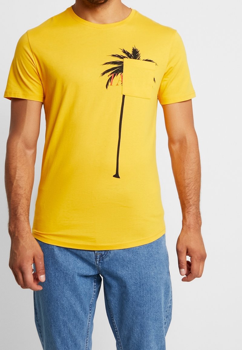 Žluté bavlněné tričko s krátkými rukávy, s potiskem černé palmy a malou kapsou na levé straně. Hladká textura.