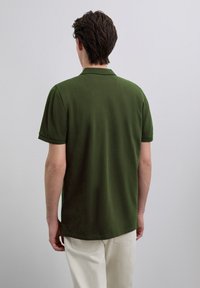 Tricou polo verde închis, fabricat din material moale. Are mâneci scurte, guler clasic și croială lejeră. Design simplu și fără cusături.
