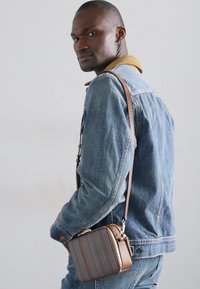 Brūna plecu soma ar teksturētu rakstu, kvadrātveida forma, sudraba aksesuāri un noņemama siksna, kas nēsāta virs zila džinsa jaka.