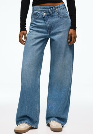 Person iført løstsiddende blå denimjeans med unik overlappende knappet taljebånd og beige sneakers, stående mod en hvid baggrund.