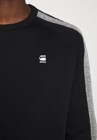 Sweat-shirt noir à col rond avec des bandes grises sur les épaules et le logo blanc "G RAW" sur la poitrine, porté par une personne.
