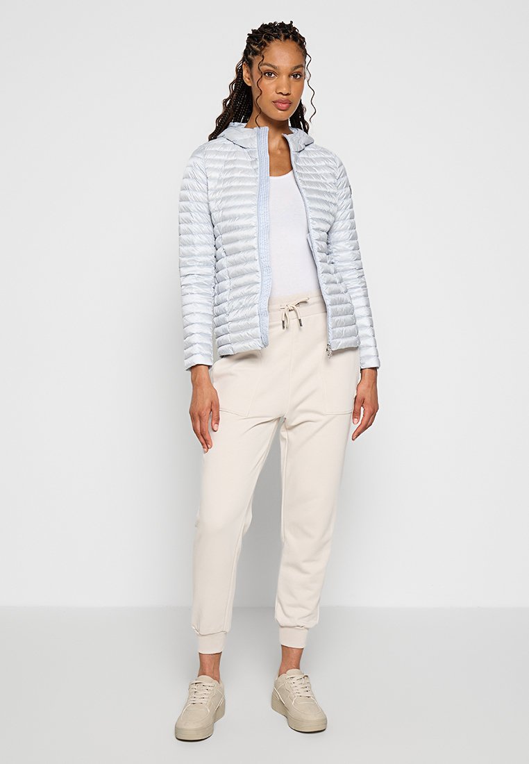 Giacca puffer azzurro chiaro con design trapuntato, chiusura con zip e cappuccio, abbinata a pantaloni beige a gamba stretta e sneaker neutre. Texture liscia.