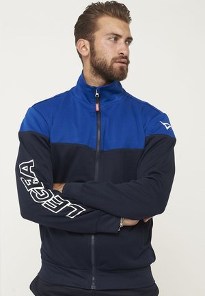 Uomo con barba che indossa una giacca blu e nera con zip con la scritta "LEGA" sulla manica destra, guarda di lato con le braccia incrociate.