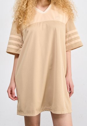 Kobieta ubrana w luźną beżową sportową sukienkę z siateczki z krótkimi rękawami i trzema paskami na każdym ramieniu, stojąca na jednolitym tle.