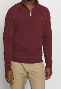 Burgundský svetr na zip s vysokým límcem, manžetami a lemem se žebrováním. Obsahuje kontrastní prošívání a malé zlaté logo na hrudi.