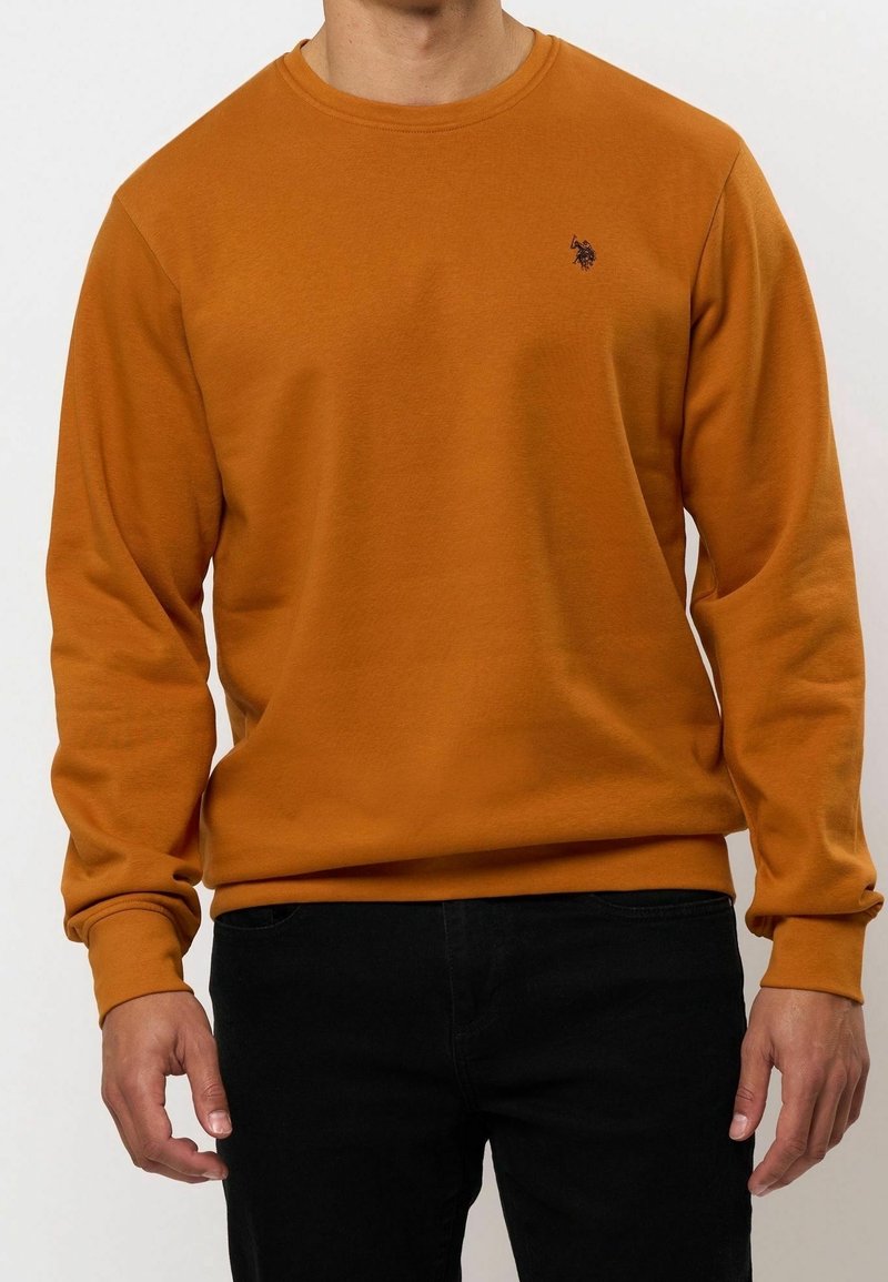 Homme portant un sweat-shirt orange uni à manches longues avec un petit logo noir brodé sur la poitrine gauche, associé à un pantalon noir.