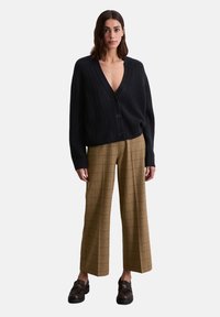 Schwarze gestrickte Karte mit tiefem V-Ausschnitt, Knopfverschluß und lockerer Passform, kombiniert mit braunen karierten weiter Beinform und schwarzen Slippern.