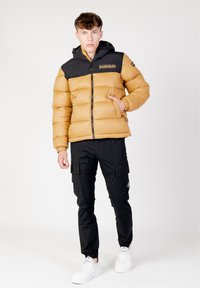 Giubbotto imbottito in beige e nero, con zip frontale, cappuccio, polsini elasticizzati e dettaglio con logo. Indossato con pantaloni cargo neri e sneakers bianche.