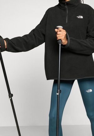 Osoba nosząca czarną bluzę polarową z zamkiem do połowy i niebieskie legginsy, trzymająca czarne kijki trekkingowe z szarymi uchwytami.
