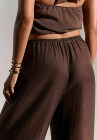Pruun linenist segu crop top ja lohvad püksid elastse vöökohtaga; sile tekstuur, kerge sära ja mugav lõige. Näha on kuldseid käevõrusid.