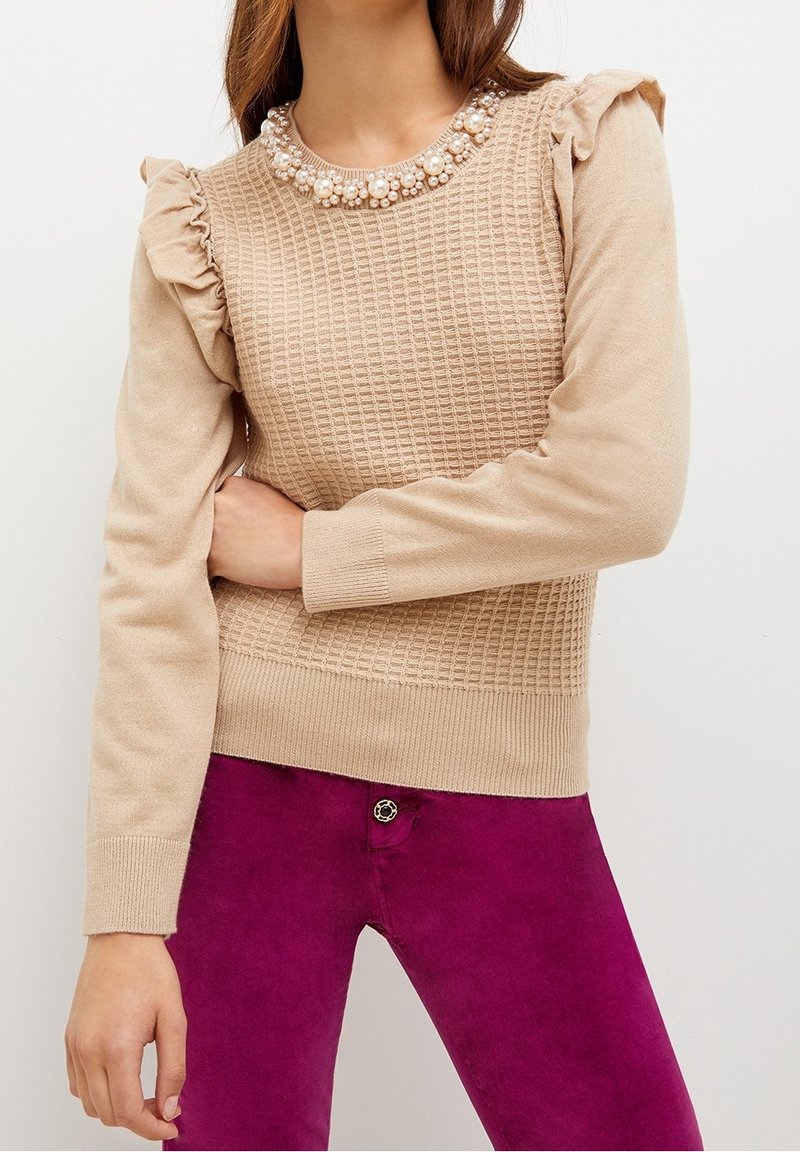 Pull en maille beige avec des manches à volants et des ornements en perles au niveau du col, associé à un pantalon violet vif.
