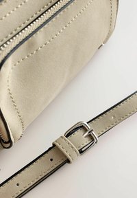 Lys beige ruskindshåndtaske med sølvlynlås og justerbar rem med sølvrektangulært spænde på hvid baggrund.