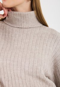 Pulover turtleneck bej deschis, cu nervuri, realizat dintr-un material tricotat moale, având un guler înalt și o siluetă ajustată.
