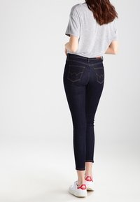 Smala jeans i mörk denim med kontrasterande sömmar. Kombineras med en grå t-shirt och vita sneakers med röda detaljer. Sedd bakifrån.