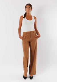 Pantaloni marroni a vita alta con gamba dritta, realizzati in tessuto liscio. Indossati con un top bianco senza maniche e décolleté neri a punta.
