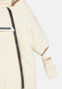 Giacca imbottita color crema con cappuccio, cerniera laterale e dettagli marroni. Presenta polsini elastici e muffole attaccate. Logo del marchio visibile sul petto.