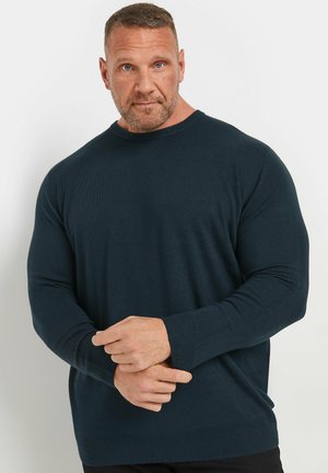 Homme musclé aux cheveux courts et à la barbe portant un pull bleu foncé à manches longues, debout devant un fond uni clair, les mains jointes devant.