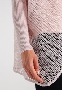 Hellrosa Strickpullover mit strukturiertem, offenem Strickdesign und einer gerippten Ärmelbündchen. Der Saum ist mit einem kontrastierenden schwarzen Mesh-Detail versehen.