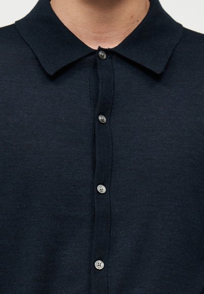 Άνδρας που φοράει σκούρο μπλε πουκάμισο με κουμπιά και μυτερό γιακά, τα κουμπιά είναι κουμπωμένα μέχρι τον λαιμό.