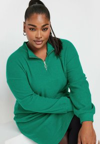 Suéter de punto verde con cremallera, textura acanalada, mangas largas y cuello alto. Presenta una cremallera plateada con un tirador circular.
