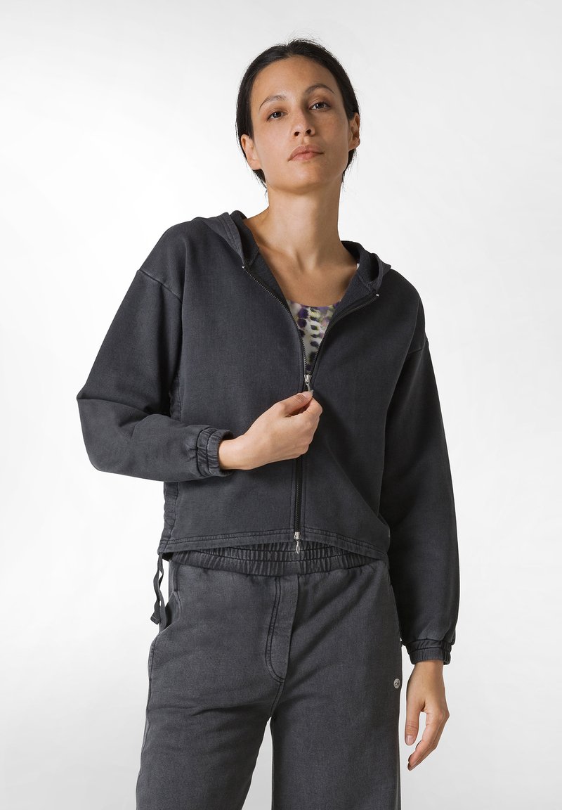 Sudadera negra con cremallera en un tejido suave, con dobladillo ajustable y puños elásticos. Combinada con pantalones deportivos negros a juego.