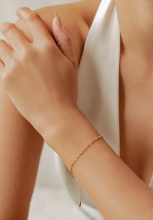 Pulsera de cadena de oro con un diseño retorcido, eslabones delicados y un cierre ajustable, llevada en una muñeca sobre un fondo claro.