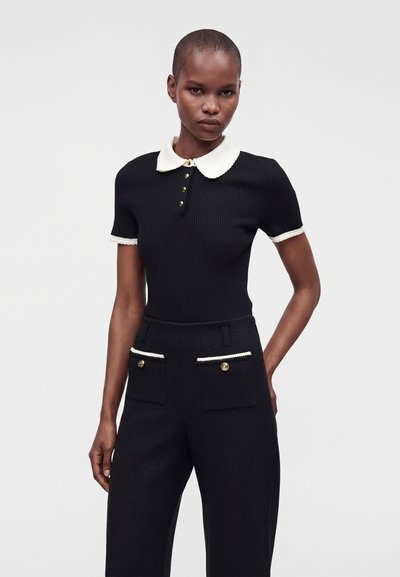 Femme portant un haut noir à manches courtes avec col et poignets blancs, associé à un pantalon noir taille haute avec poches avant boutonnées.