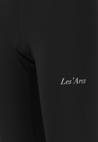 Mustat leggingsit, joissa on sileä pinta, ja alhaalla reidessä valkoinen "Les'Arcs" -logo sekä vahvistetut sivusaumat.
